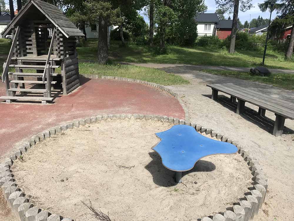 Sandlåda med blå sittplats. Klätterträd med koja i bakgrunden. Sittbänkar