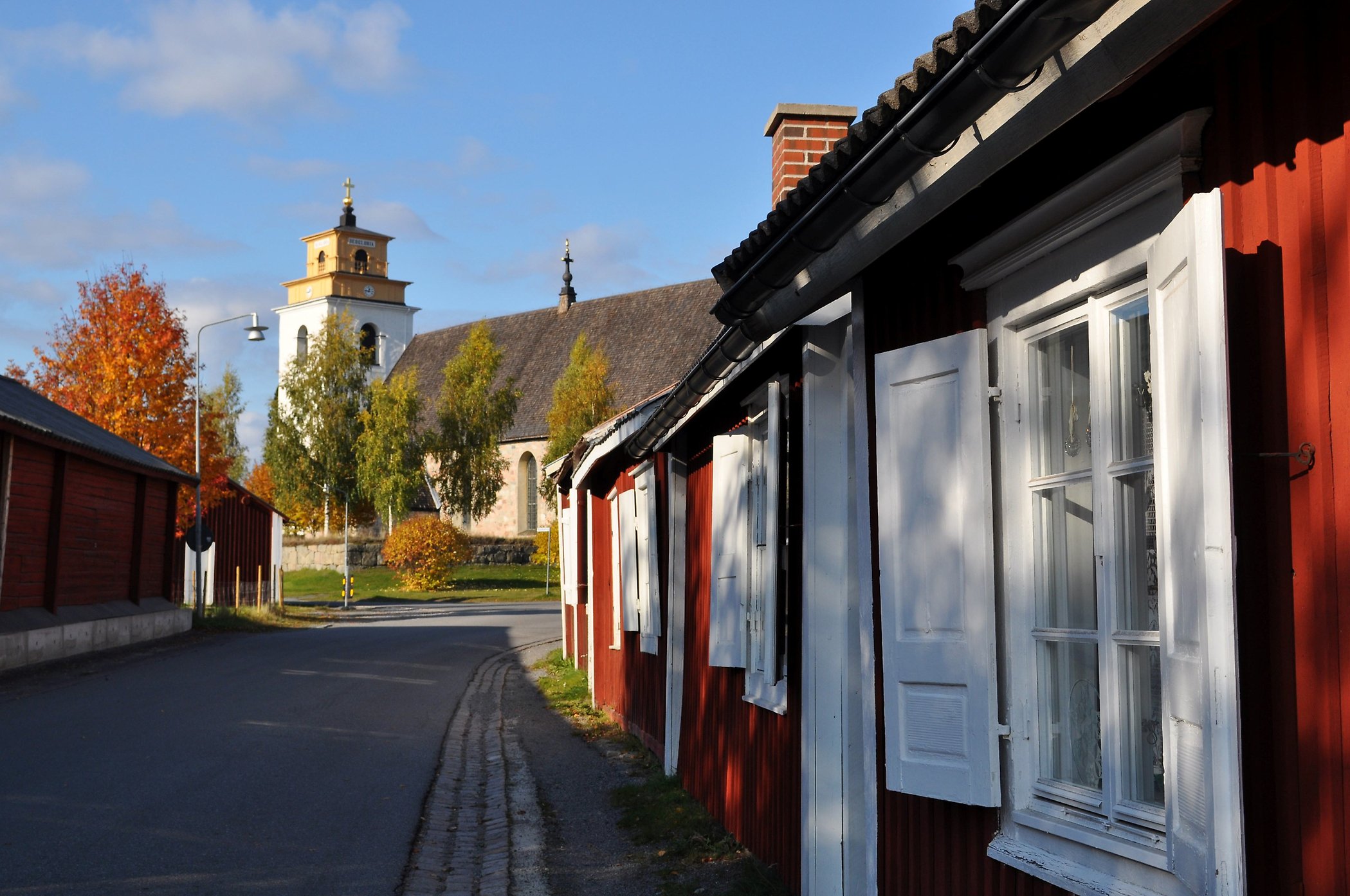 Väg med små röda och vita kyrkstugor i Gammelstads kyrkstad. Kyrka och höstträd i bakgrunden.