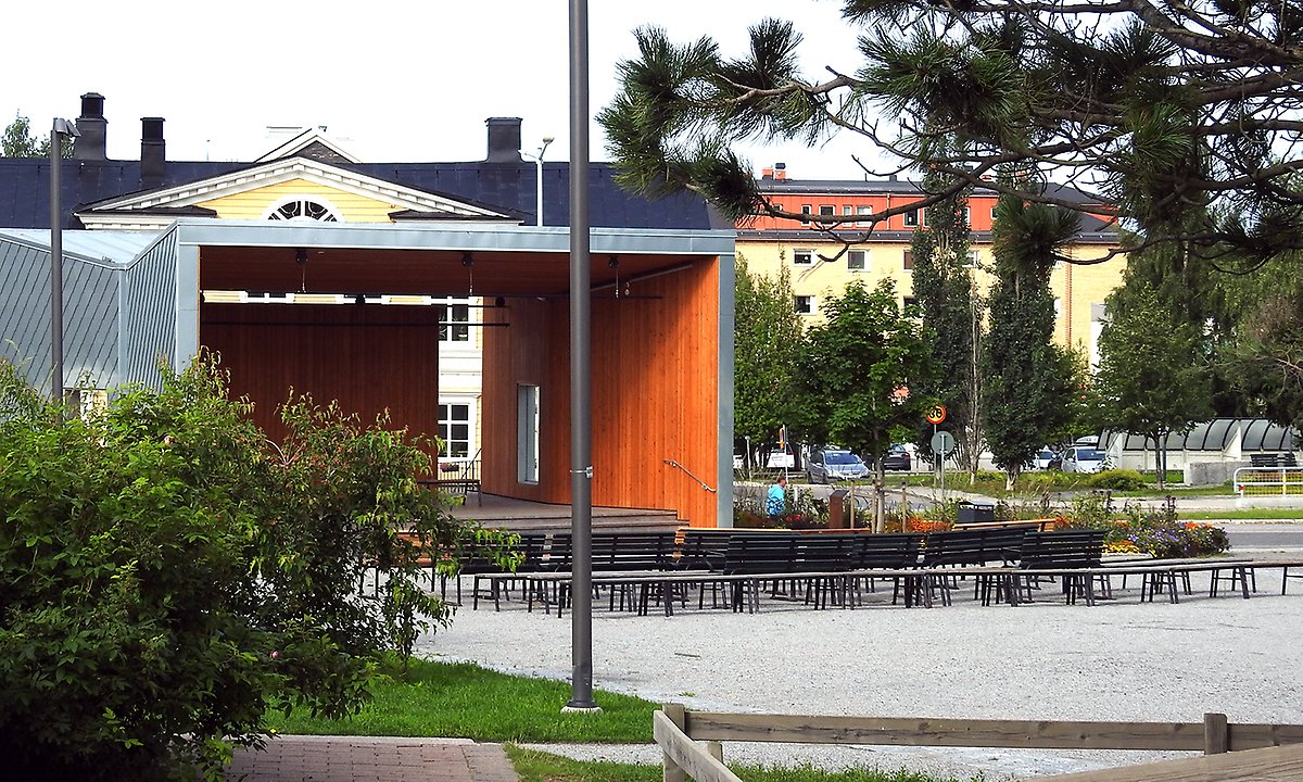 Stadsparken väg mot scenen med träd i förgrunden. Belysningsstolpar är riktade mot träden